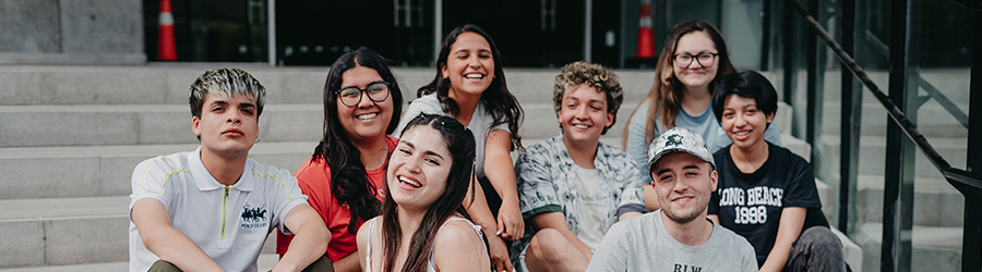 En la foto aparece un grupo de estudiantes felices sentadas(os) en la escalera del frontis de la Facultad de Administración y Economía