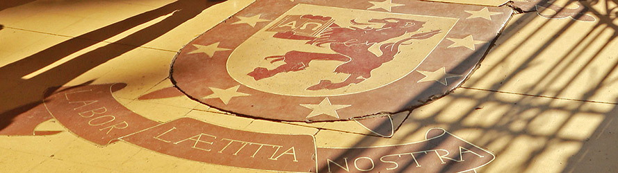 En la imagen aparece el escudo de la universidad ubicado en el piso de la entrada a la Escuela de Artes y Oficios, con el texto "Labor laetitia nostra" en la parte inferior de él.